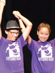 Two kids in purple "Studio East" T-shirts, one wearing glasses and a hat, holding hands with arms raised in a celebratory manner.
