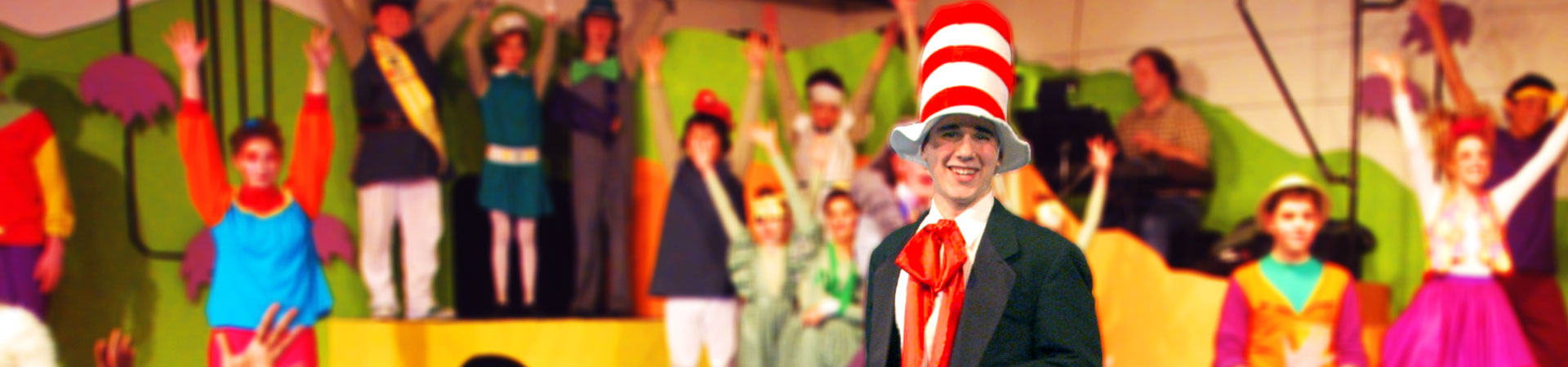 Seussical, the Musical is performed live at Studio East Training for the Performing Arts in Kirkland, WA. The Cat in the Hat is the focus. In the background, many young performers dressed in bright Whovian costumes raise their hands to the sky.